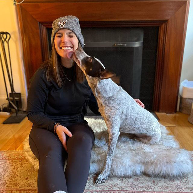 Young woman sitting on the floor wearing an oxford grey custom dog beanie with her dog's face on it while her dog licks her cheek.